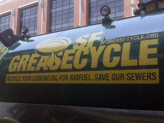 A green truck with “SF Greasecycle” and “Recycle your cooking oil for biofuel. Save our sewers” printed on the side, highlighting grease trap recycling, is parked in front of a brick building.
