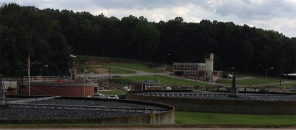 A wastewater treatment facility featuring large circular processing tanks with integrated grease trap systems in the foreground, administrative buildings in the background, and surrounded by trees and grassy areas under a cloudy sky.