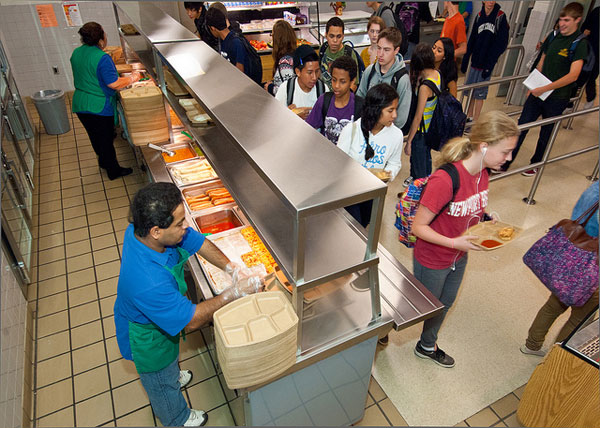 A cafeteria worker serves food to a line of students waiting with trays in a school cafeteria. The students, wearing backpacks and casual clothes, choose from meals displayed behind a sneeze guard near the kitchen’s grease trap.