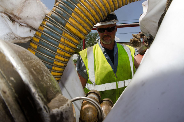 A worker wearing a yellow safety vest, helmet, and sunglasses stands behind large industrial hoses and pipes near a grease trap outdoors on a sunny day.