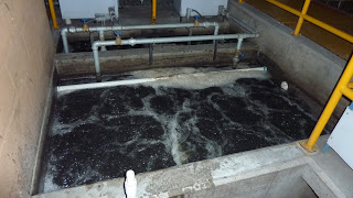 A rectangular concrete tank, likely a grease trap, filled with dark, bubbling water and surrounded by pipes, railings, and equipment—typical of an industrial or wastewater treatment facility.