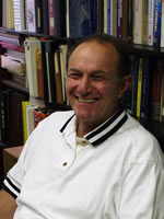 A man wearing a white polo shirt with black stripes on the collar is smiling while seated in front of a bookshelf filled with books and binders, possibly discussing topics like maintenance or grease trap solutions.